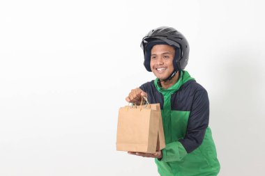 Yeşil ceket ve kask giyen Asyalı kurye sürücünün portresi içinde hediye olan el işi kağıt alışveriş torbası veriyor. Gri arkaplanda izole edilmiş resim