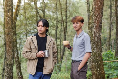 Ahşap ve paslanmaz çelik bardakla birlikte içerken ormanın tadını çıkaran iki Asyalı adamın portresi. Dağ yürüyüşü konsepti