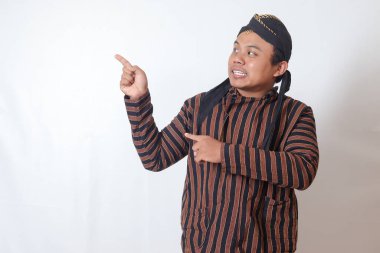 Boş uzayı parmakla gösteren beyaz şapkalı, java kumaşlı heyecanlı Asyalı adamın portresi. Reklam ve tanıtım konsepti. Beyaz arkaplanda izole edilmiş resim