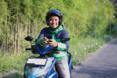 Online taxi rider wearing green jacket and helmet, riding motorcycle, using a mobile phone while on the way to pick up customer. Transportation application concept. clipart