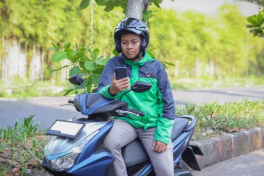 Online taxi rider wearing green jacket and helmet, riding motorcycle, using a mobile phone while on the way to pick up customer. Transportation application concept. clipart