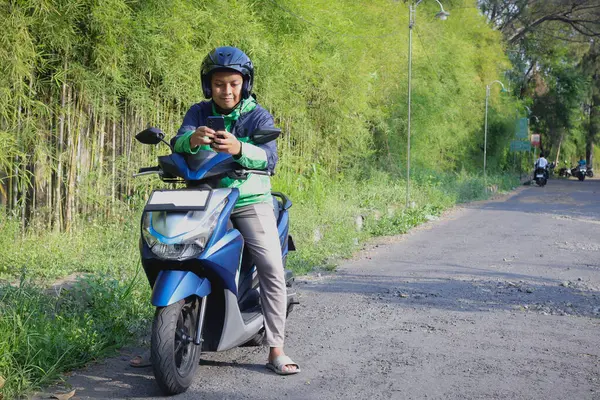 Çevrimiçi taksi şoförü yeşil ceket ve kask giyiyor, motosiklet sürüyor, müşteri almaya giderken cep telefonu kullanıyor. Ulaşım uygulaması kavramı.
