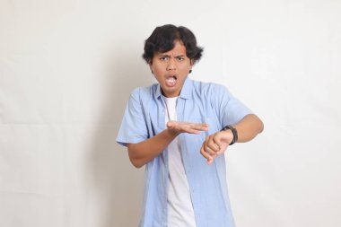 Mavi gömlekli heyecanlı Asyalı adamın portresi kol saatine bakıyor. Reklam konsepti. Beyaz arkaplanda izole edilmiş resim