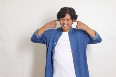 Sevimsiz Asyalı bir adamın portresi mavi gömlekli gözlerini kapatan, kulaklarını parmaklarıyla kapatan. Beyaz arkaplanda izole edilmiş resim