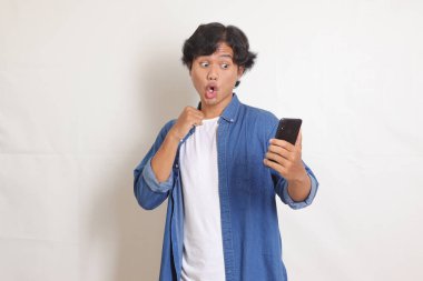 Heyecanlı mavi gömlekli Asyalı adamın portresi cep telefonu kullanırken yumruğunu kaldırıp başarıyı kutluyor. Beyaz arkaplanda izole edilmiş resim