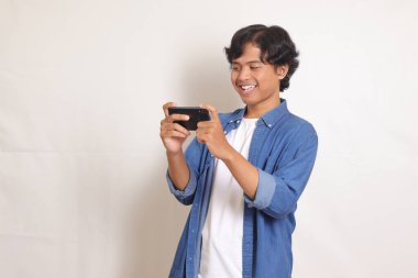 Mavi gömlekli heyecanlı Asyalı adamın portresi ekranı eğerek cep telefonuyla oyun oynuyor. Vay be yüz ifadesi. Beyaz arkaplanda izole edilmiş resim