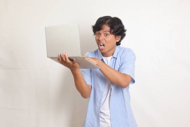 Mavi gömlekli kafası karışık Asyalı bir adamın portresi dizüstü bilgisayarını tutarken elini yayıyor. Reklam konsepti. Beyaz arkaplanda izole edilmiş resim