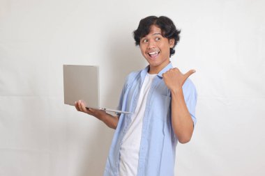 Mavili tişörtlü heyecanlı Asyalı adamın portresi. Elinde dizüstü bilgisayarla ürünü gösteriyor ve ona doğrultuyor. Beyaz arkaplanda izole edilmiş resim