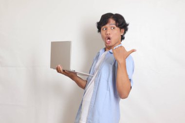 Mavili tişörtlü heyecanlı Asyalı adamın portresi. Elinde dizüstü bilgisayarla ürünü gösteriyor ve ona doğrultuyor. Beyaz arkaplanda izole edilmiş resim