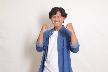 Mavi gömlekli çekici Asyalı adamın portresi yumruğunu kaldırarak başarıyı kutluyor. Beyaz arkaplanda izole edilmiş resim