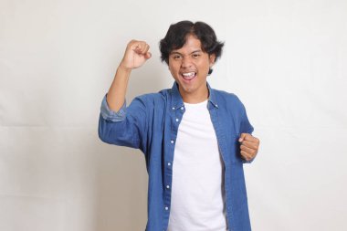 Mavi gömlekli çekici Asyalı adamın portresi yumruğunu kaldırarak başarıyı kutluyor. Beyaz arkaplanda izole edilmiş resim