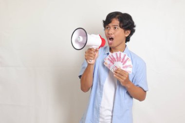 Mavi gömlekli heyecanlı Asyalı adamın portresi megafon kullanarak konuşuyor, rupiah para banknotunu tutarken ürünü tanıtıyor. Reklam konsepti. Beyaz arkaplanda izole resimler