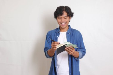 Mavi gömlekli çekici Asyalı adamın portresi rupiah para banknotunu tutarken kişisel finans hakkında notlar yazıyor. Finansal eğitim kavramı. Beyaz arkaplanda izole resimler