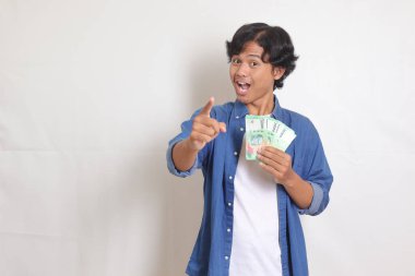Mavi gömlekli çekici Asyalı adamın portresi rupiah para banknotu tutuyor ve parmağıyla işaret ediyor. Beyaz arkaplanda izole resimler