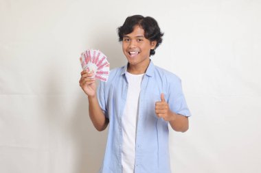 Portrait of attractive Asian man in blue shirt holding rupiah money banknote and pointing side with finger. Isolated images on white background clipart