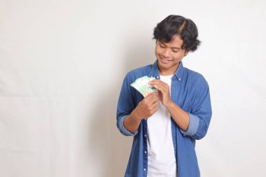 Mavi gömlekli çekici Asyalı adamın portresi rupiah para banknotu tutuyor ve heyecanlanıyor. Beyaz arkaplanda izole resimler
