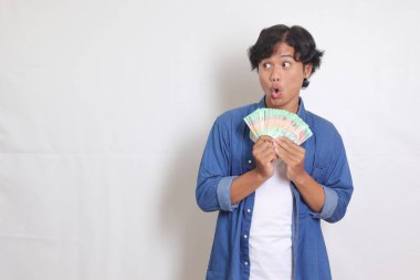 Mavi gömlekli çekici Asyalı adamın portresi rupiah para banknotu tutuyor ve heyecanlanıyor. Beyaz arkaplanda izole resimler