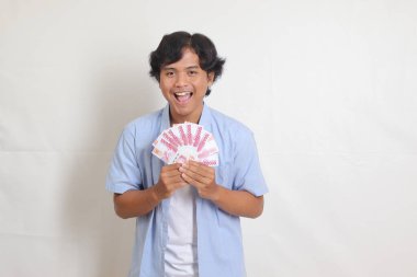 Mavi gömlekli çekici Asyalı adamın portresi rupiah para banknotu tutuyor ve heyecanlanıyor. Beyaz arkaplanda izole resimler