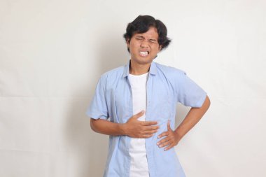 Mavi gömlekli, midesinde ağrı hisseden yorgun Asyalı bir adamın portresi. Açlığa katlanıyor. Karnım ağrıyor ve yemek konseptine açım. Beyaz arkaplanda izole edilmiş resim