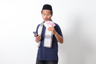 Sarung eşarp takan Asyalı Müslüman adamın portresi elinde rupiah para banknotuyla cep telefonuna bakıyor. Beyaz arkaplanda izole edilmiş resim