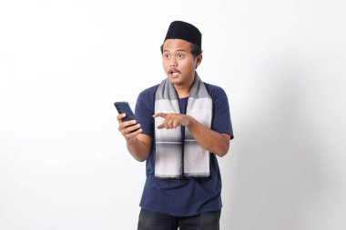 Portrait of upset Asian muslim man wearing sarung scarf and skullcap, holding mobile phone and showing shocked expression. Isolated on white background clipart