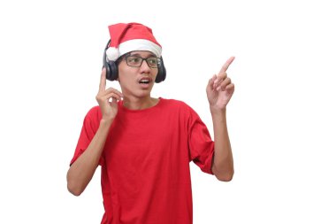 Kırmızı tişörtlü, Noel Baba şapkalı heyecanlı Asyalı adamın portresi kulaklık kullanarak müzik dinliyor ve eğleniyor. Beyaz arkaplanda izole edilmiş resim