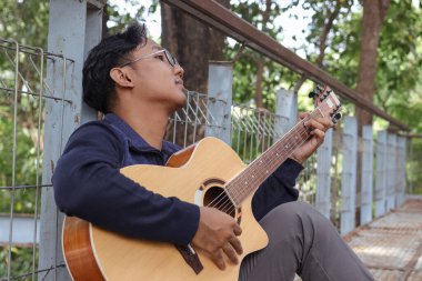 Yaz boyunca bir parkta gitar çalan Asyalı bir adamın portresi, yemyeşil ve mevsimin huzurlu atmosferiyle çevrili.