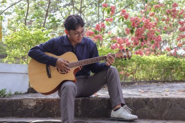 Asyalı bir adam sıcak bir yaz gününde yeşil alanlarla çevrili şehir parkında gitar çalar. Huzurlu atmosfer, parlak güneş ışığı ve rahatlatıcı melodi huzurlu ve enerjik bir an yaratır.