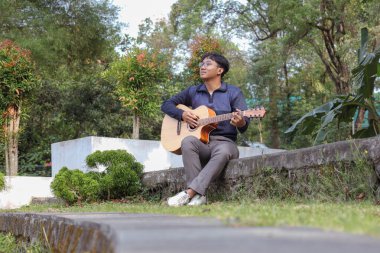 Asyalı bir adam sıcak bir yaz gününde yeşil alanlarla çevrili şehir parkında gitar çalar. Huzurlu atmosfer, parlak güneş ışığı ve rahatlatıcı melodi huzurlu ve enerjik bir an yaratır.