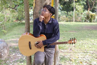 Asyalı bir adam sıcak bir yaz gününde yeşil alanlarla çevrili şehir parkında gitar çalar. Huzurlu atmosfer, parlak güneş ışığı ve rahatlatıcı melodi huzurlu ve enerjik bir an yaratır.