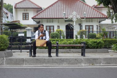 Beyaz tişörtlü Asyalı iş arayanın portresi özgeçmişini içeren kahverengi bir zarf tutuyor, akıllı telefon kullanıyor, başvurduğu şirketten gelen e-postaları ve haberleri kontrol ediyor ve bankta oturuyor.