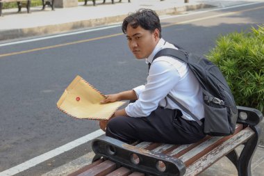 Beyaz gömlekli Asyalı iş arayanın portresi. Özgeçmişini içeren kahverengi bir zarf tutarken iş arıyor. Bankta oturuyor. Personel alımı konsepti