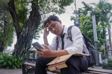 Beyaz tişörtlü Asyalı iş arayanın portresi özgeçmişini içeren kahverengi bir zarf tutuyor, akıllı telefon kullanıyor, başvurduğu şirketten gelen e-postaları ve haberleri kontrol ediyor ve bankta oturuyor.