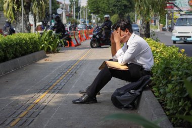 Takım elbiseli Asyalı bir çalışanın portresi. Elinde özgeçmişi olan kahverengi bir zarf var. Kaldırımda elleri başının üstünde oturuyor. İşten çıkarma ve işsizlik kavramı