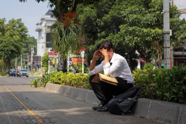 Takım elbiseli Asyalı bir çalışanın portresi. Elinde özgeçmişi olan kahverengi bir zarf var. Kaldırımda elleri başının üstünde oturuyor. İşten çıkarma ve işsizlik kavramı
