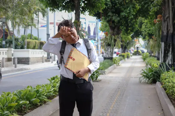 Özgeçmişini içeren kahverengi bir zarf taşırken terini silen takım elbiseli Asyalı bir iş arayanın portresi. Personel alımı konsepti.