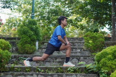 Asyalı bir adam sabah koşusundan önce şehir parkında bacaklarını esnetiyor, ısınma egzersizine hazırlanıyor.