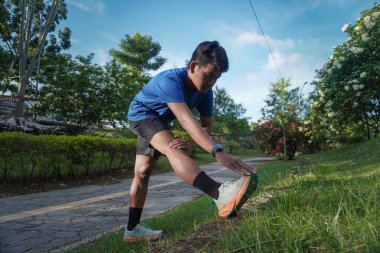 Asyalı bir adam sabah koşusundan önce şehir parkında bacaklarını esnetiyor, ısınma egzersizine hazırlanıyor.