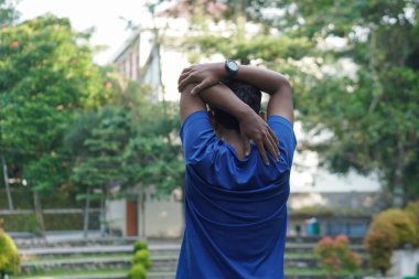 Asyalı bir adam sabah koşusundan önce şehir parkında kollarını uzatıyor, ısınma egzersizine hazırlanıyor.