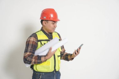 Güvenlik yeleği ve miğfer takan Asyalı inşaat işçisi, elinde proje kağıdı tutarken bir telefon planlıyor, internet sitesini yönetirken konuşuyor.