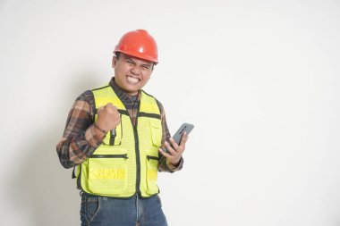 Asyalı inşaat işçisi, güvenlik yeleği ve kask takmış, akıllı telefonu tutarken mutlu bir şekilde gülümsüyor, heyecan ve pozitif enerji gösteriyor. Beyaz arkaplanda izole edilmiş resim