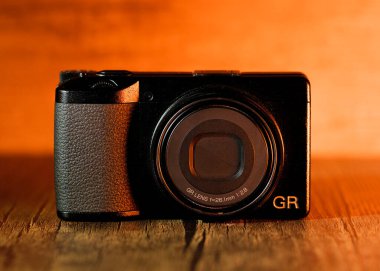 Ricoh GR III x, ahşap arka planda serin turuncu ışıkları olan 24MP APS-C format kamerasıdır.