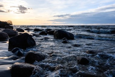 Kıyıdaki granit taşların üzerinde köpüren deniz dalgaları. Bulutlu sis güneşi biraz gizliyor. Gün batımından bir saat önce. Arka ışık. Narva Körfezi, Baltık Denizi, Estonya