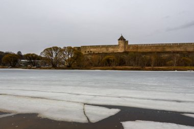 Nehrin bahar manzarası dondurucu soğuğun sonunda ve buz kaymasının başlangıcında. Arka planda filizlenen tomurcukları ve ortaçağ kalesi olan çalılar ve söğüt ağaçları var. Bulutlu gün