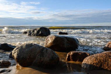 Köpüklü deniz dalgaları kıyıdaki granit taşlara hücum ediyor. Alçak akşam güneşinden direkt ışık. Taşlar gölgeler bırakır. Mavi gökyüzü hafif bulutlu ve puslu. Ufukta gün batımı pembeleri beliriyor