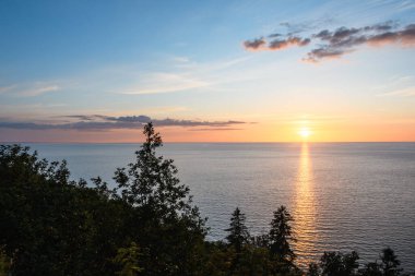 Baltık Denizi üzerinde gün batımı ve ormanlık sahil. Akşam mavisi gökyüzü ve güzel bulutlar kırmızı ve sarı renkli. Güneşli yol, ufuktan sahile doğru bir parıltı. Üçgenlerle bileşim