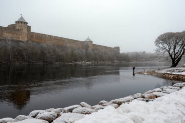 Kış, donmuş bir nehir eski bir kale arasında akar (15 sent). Ve karla kaplı modern granit seti. Elinde olta olan bir balıkçı kıyıya yakın sularda duruyor.