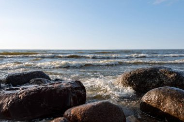 Köpüren deniz dalgaları ön ve ritmik olarak büyük pembe granit taşların üzerine yuvarlanır. Güneşli bir yaz akşamında mavi gökyüzü. Deniz manzarasında bir esinti var.