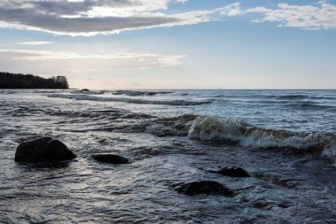Bulutlu bir gökyüzünün altındaki kayalık sahilde dalgalar. Deniz dalgalarının taşlara çarptığı doğa sahnesi. Kıyı manzarası.