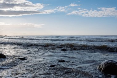 Dalgalı deniz, karanlık su ve dağılmış kayalar parçalı bulutlu bir gökyüzünün altında. Doğal dokuları ve hareket hissi olan kıyı manzarası..